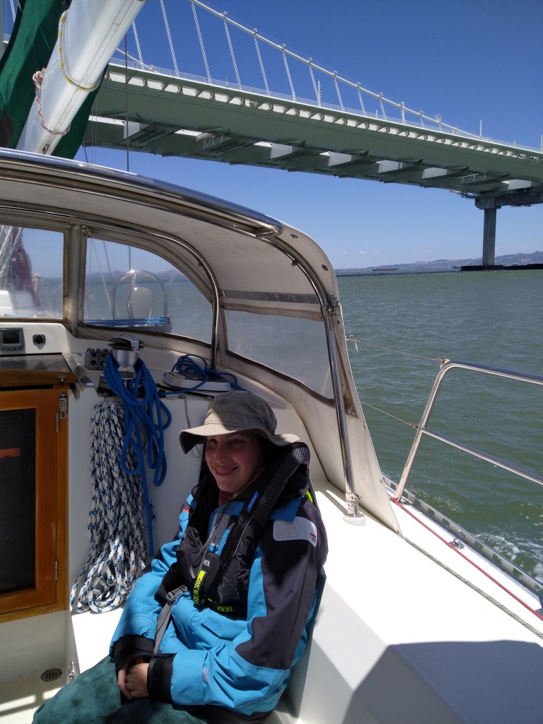 Tiffany sits in Luna Moth's cockpit wearing a sailing jacket, inflatable PFD, and a wide brimmed hat as the boat motors under the Oakland span of the San Francisco Bay Bridge.