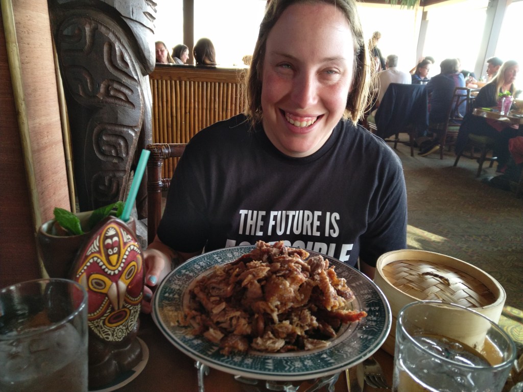 Tiffany, wearing a black t-shirt reading "The Future is Accessible" sits smiling at a restaurant table, a massive plate of shredded duck, flanked by a steaming basket and a large tropic drink, sit before her. Over her right shoulder is a large tiki statue.