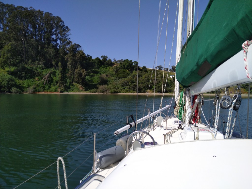 A view from the cockpit of a large sailboat shows a quiet anchorage bordered by a sand beach and a steep wooded hill.