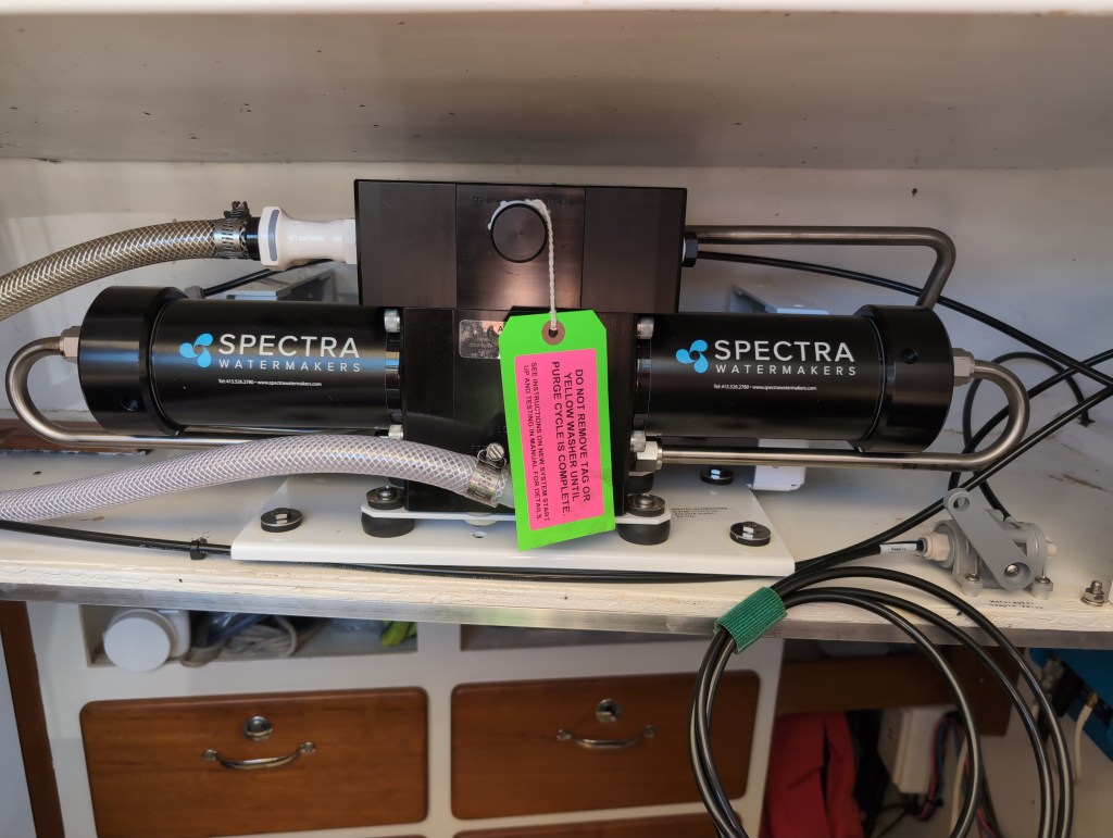 A complex assembly of a thick black cylinder and a black box with metal and plastic tubing coming out of it sits on a re-enforced wooden shelf.