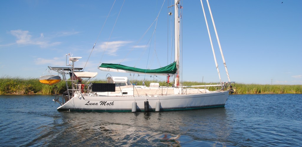A white sailboat with a yellow dinghy motors up a narrow slough with reads growing on the far bank.