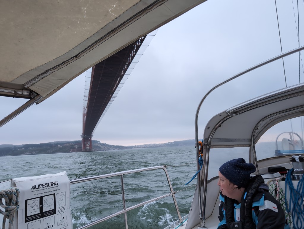 Tiffany, wearing a blue sailing jacket and PFD, looks out as the boat passes under the Golden Gate Bridge.