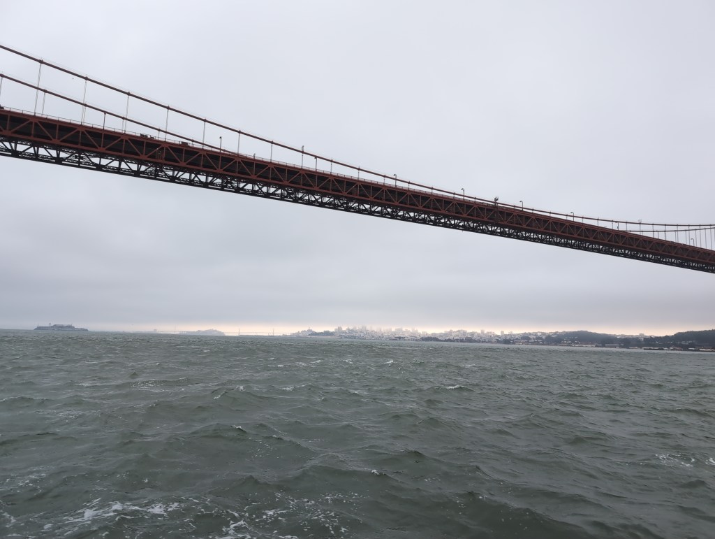 The view shows the span of the Golden Gate Bridge, with choppy water beneath it and city of San Francisco beyond. Low clouds hang over the bay, which includes the island of Alcatraz.