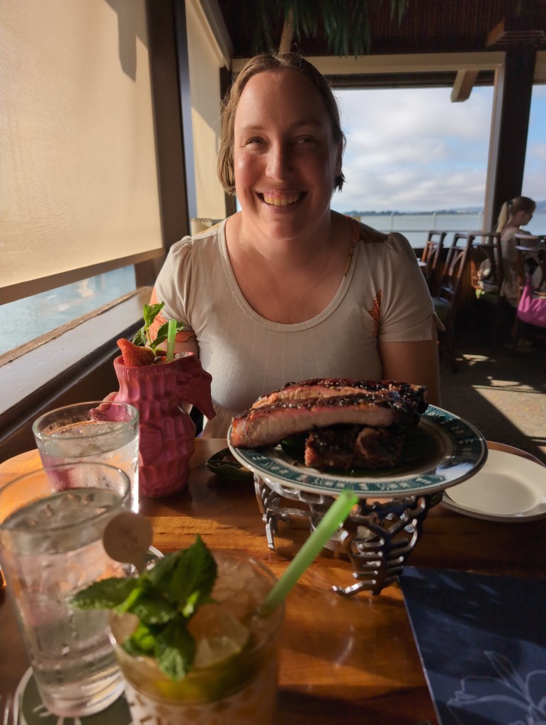 Tiffany sits at a side table, grinning, with an appetizer plate of ribs and a pink seahorse shaped cockpit glass in front of her. Out the window behind her is a view of the bay.