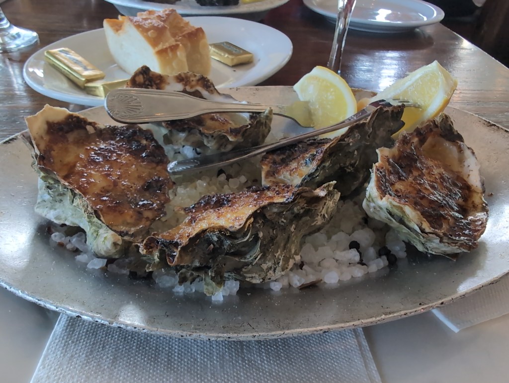 Five baked oysters sit on a bed of salt, adorned with two fork skewered lemon wedges.