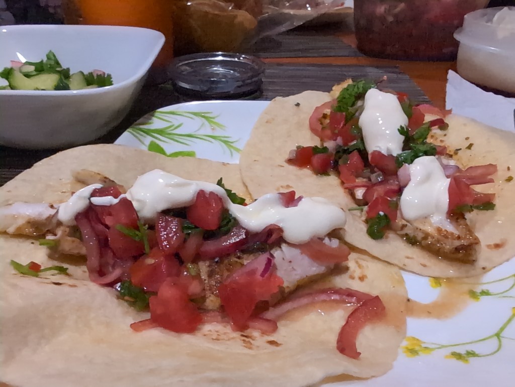A white dinner plate holds two fish tacos with salsa, marinated red onions, and crema. Behind the plate is a bowl containing cucumber salad.