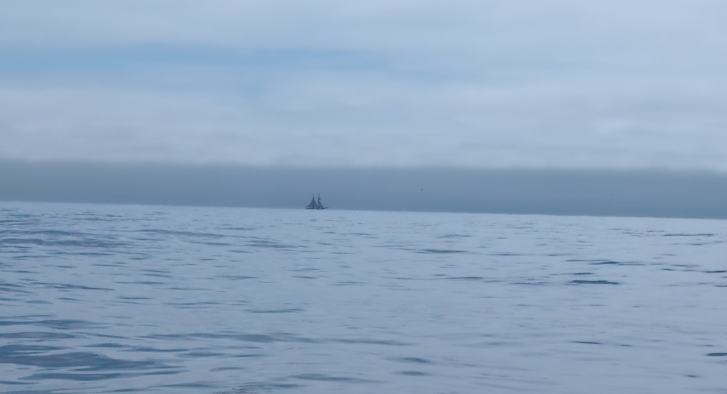 A two masted sailing vessel is visible on the horizon, over a calm sea.