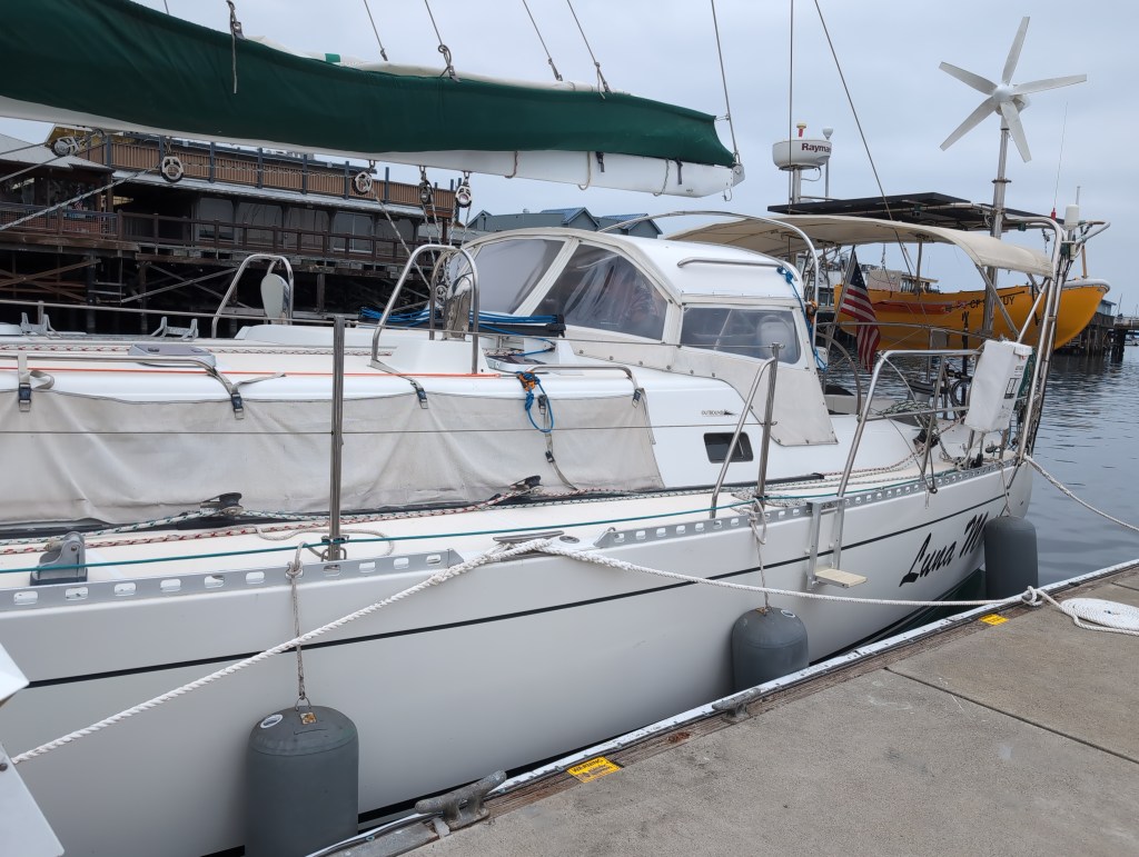 A white sailboat is sidetied to a cement dock. Across the fairway behind her is a pier with stores and restaurants.