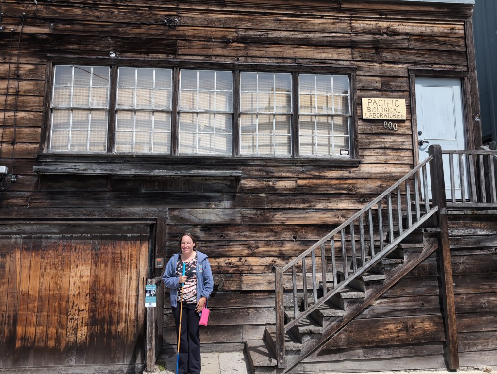 Tiffany, wearing a grey hoodie and holding a yellow and blue cane, stands in front of a weathered wooden building. A plaque by the second story entrance reads "Pacific Biological Laboratories".