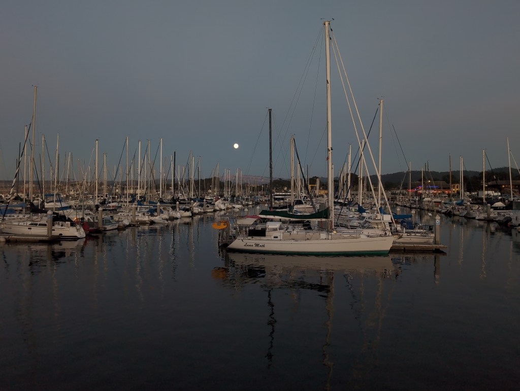 Luna Moth is seen in the evening, tied up on the end of a dock in a busy marina. A blood moon is in the sky over distant hills.