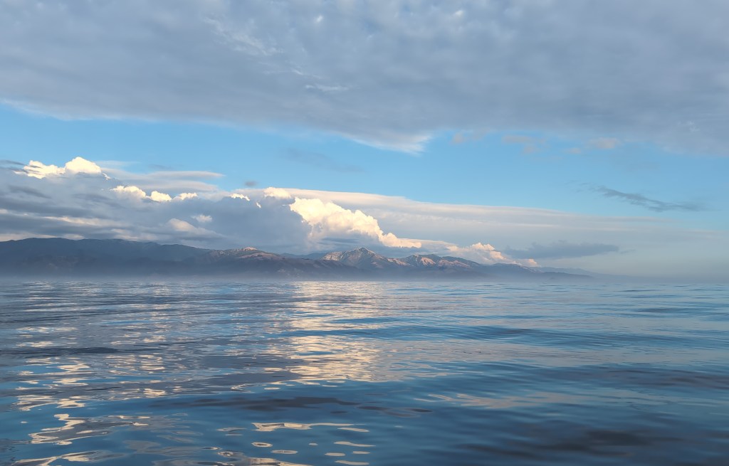 A coastal range is visible in the distance across calm seas. Low clouds hang over the hills and are reflected in the smooth surface of the water.