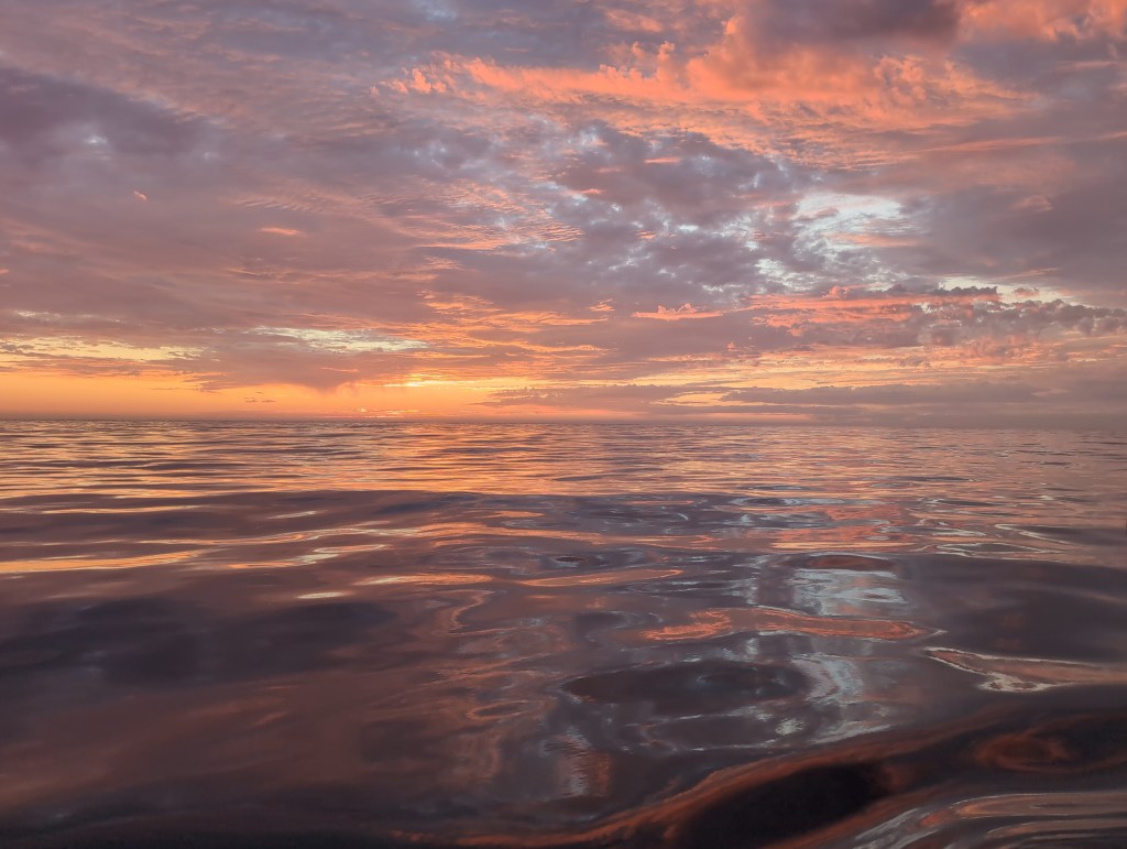 Open ocean under a vibrant sunset. The sky is filled with clouds illuminated in soft pink, orange, and purple hues, reflecting across the calm, glassy surface of the water. The horizon is visible in the distance, blending seamlessly between the sky and sea. The water appears almost mirror-like, beautifully capturing the warm colors of the sky above. There are no landforms, boats, or people in sight—just the tranquil, expansive seas and the glowing evening sky.