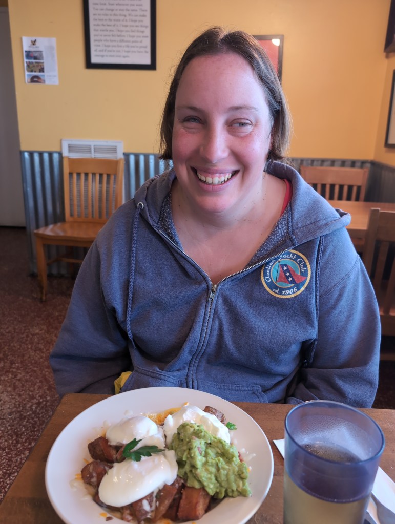 Tiffany, wearing an Aeolian Yacht Club hoodie, sits smiling in a restaurant. a plate of potatoes, poached eggs, and guacamole before her.