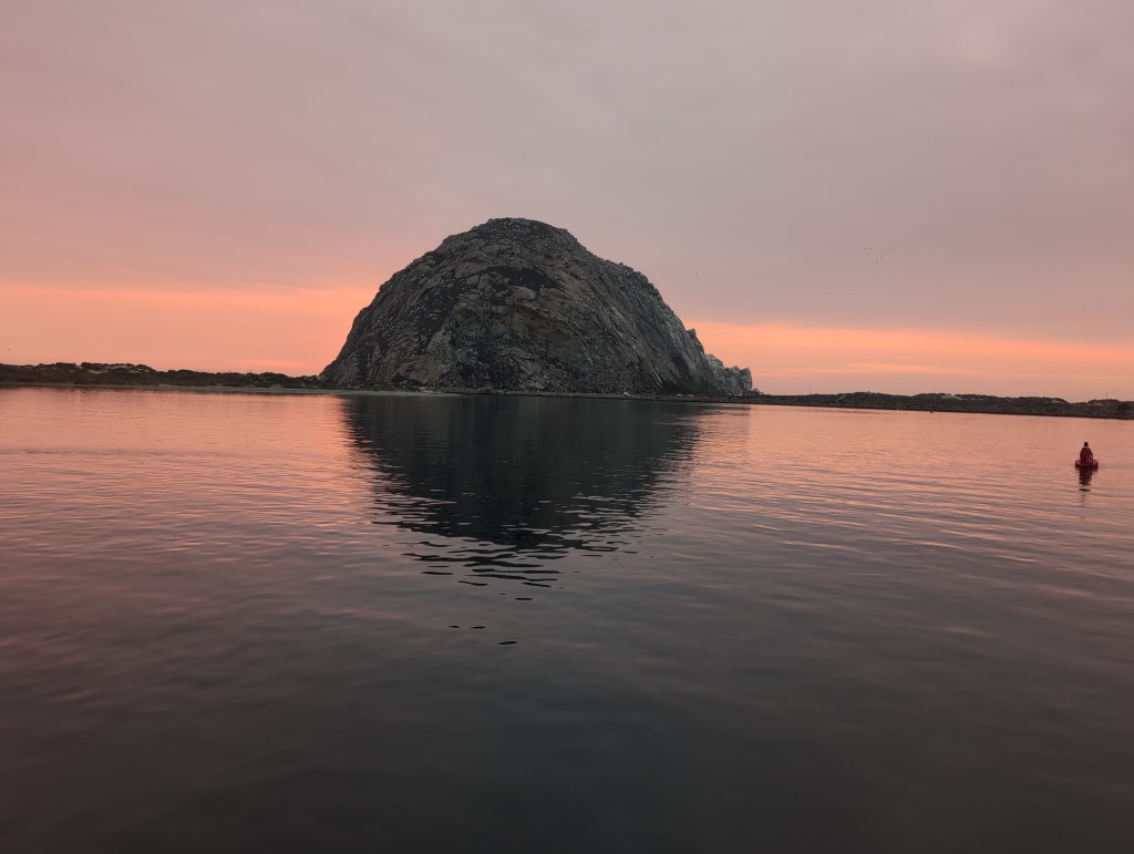 A large, dome shaped rock formation is seen across calm water with a pink sunset showing on low hanging clouds. The water reflects the color of the sunset in its smooth surface.