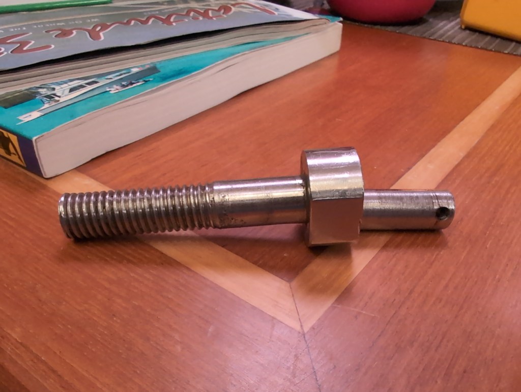 A long, thick, stainless steel pin with threads on one end, and a thick portion in the middle, lies in two pieces on a wooden table.