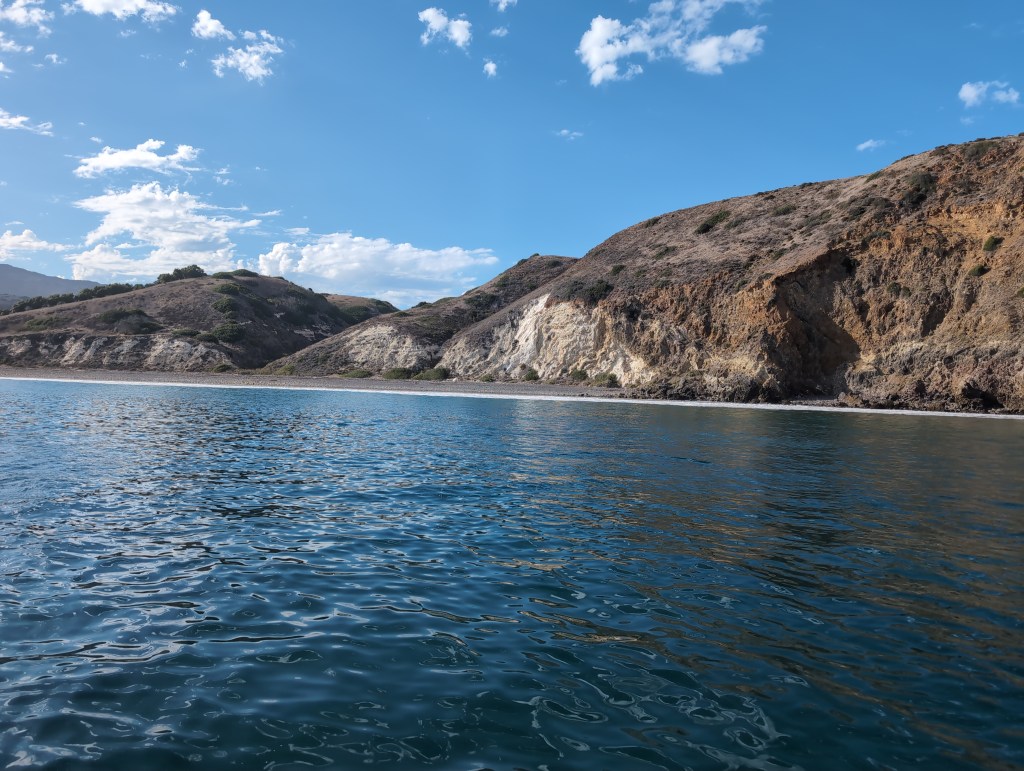 Waves break on a rocky beach in front of hills dotted with low vegetation. A white cliff shows signs of past island erosion.