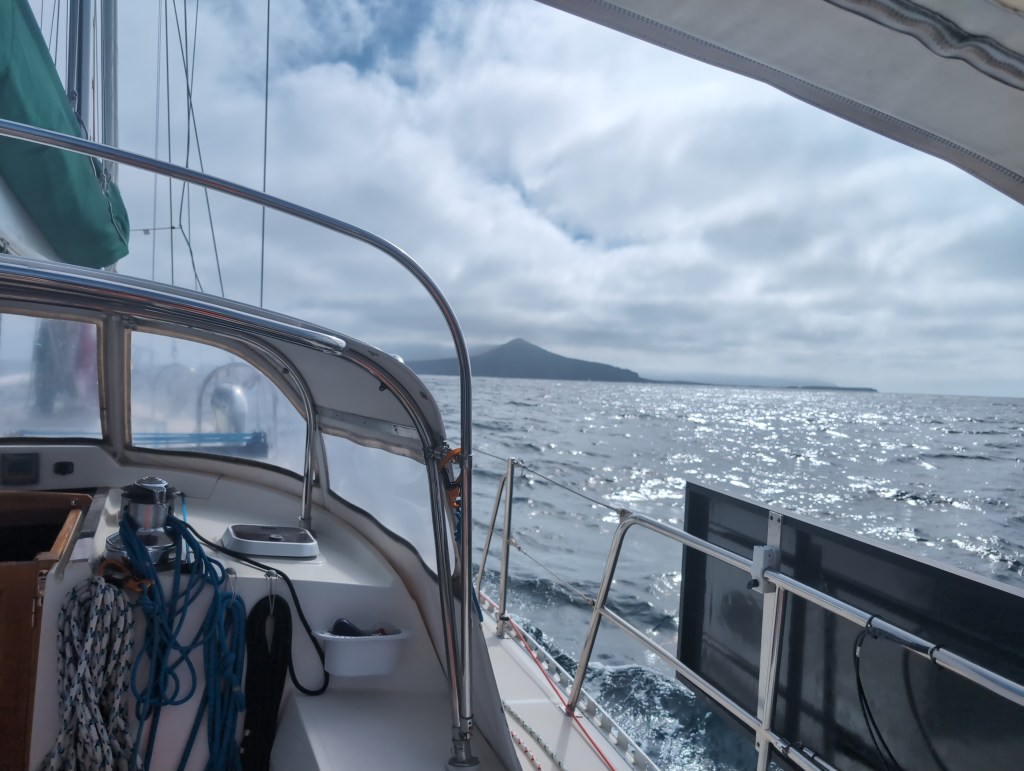A sailboat cross calm seas under an overcast sky.