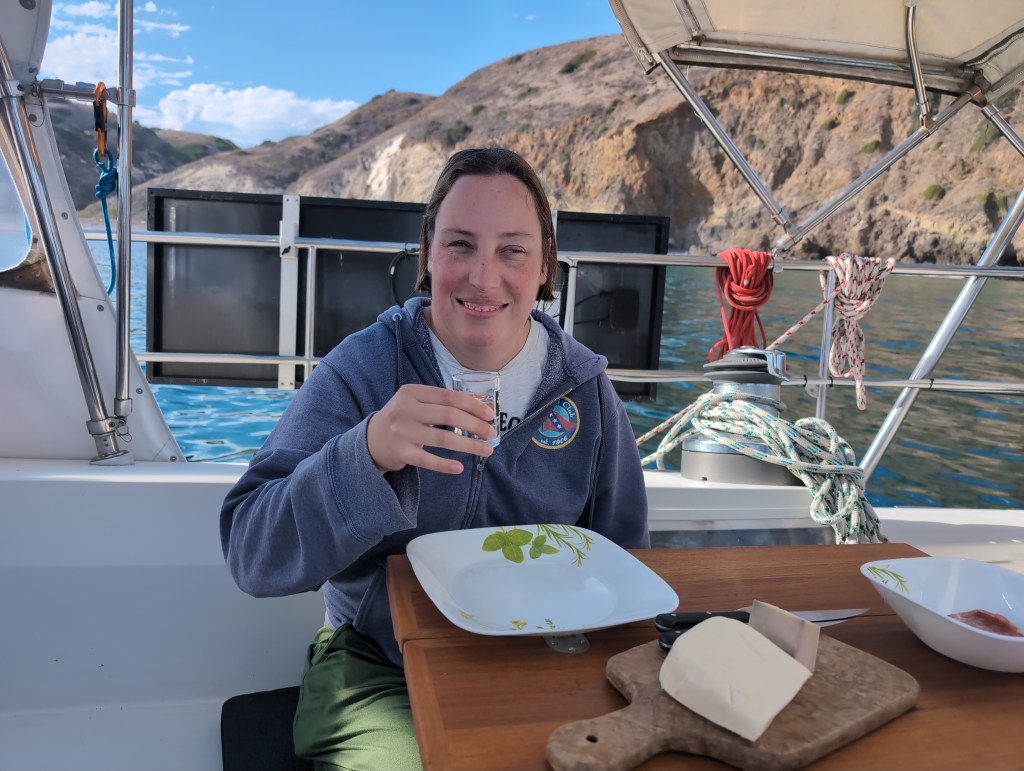 Tiffany, sitting in Luna Moth's cockpit with a view of cliffs behind her, smiles as she raises a shot glass in a toast. Before her is a wooden table with plates and a cheese board.
