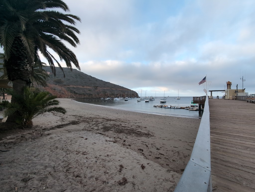 A sand beach leads to a calm harbor with a couple of dozen boats on moorings. Low clouds hang over the scene and a flag shows a slight breeze.