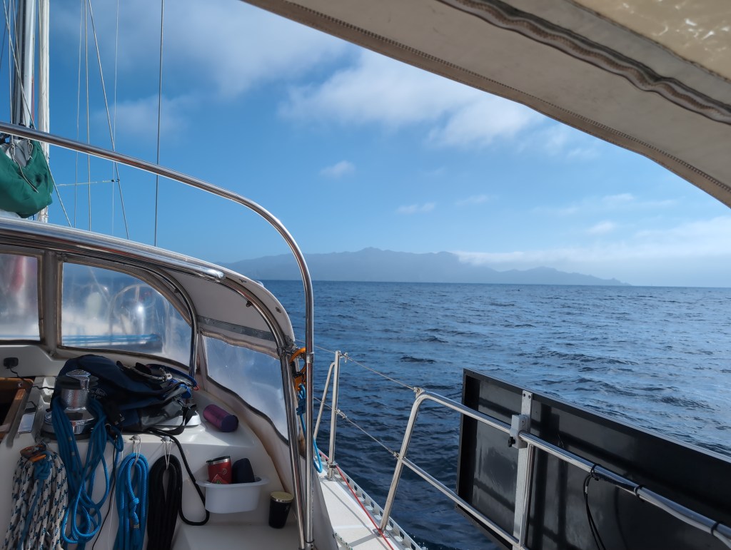 The view from the cockpit of a sailboat as it heads across calm water toward a distant island.