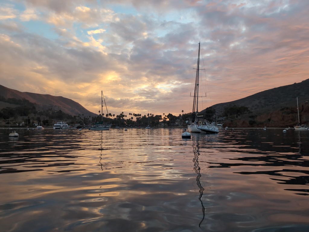 Calm water in a harbor reflects a sunset of pinks and yellows beneath low hanging clouds.