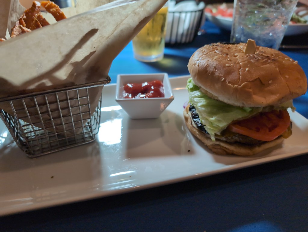 A rectangular, white plate holds a burger piled high with produce and a paper lined basket of sweet potato fries. 