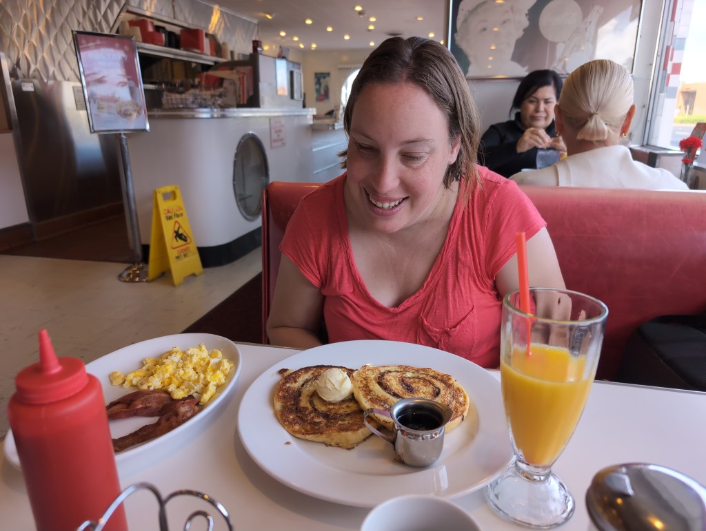 TIffany, sitting in a 50's style diner, looks down at a plate of cinnamon swirl French toast with a side of scrambles eggs and bacon. 