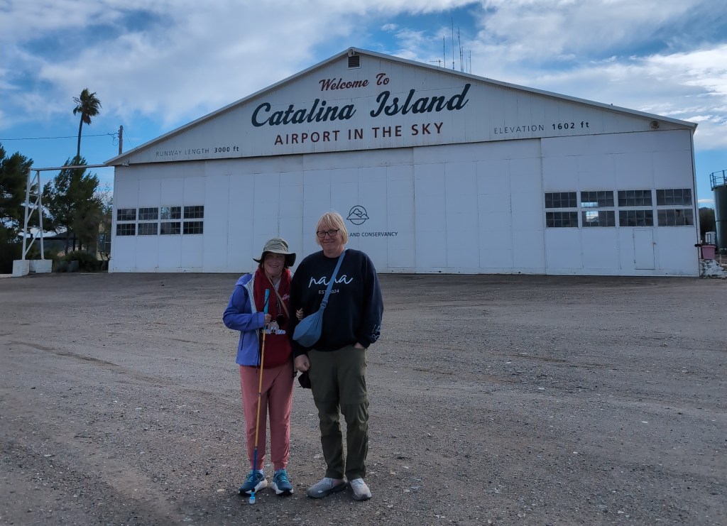 Tiffany and Lisa pose for a picture in front of a white hanger building labeled "Welcome to Catalina Island Airport in the Sky, Elevation 1602 ft" 