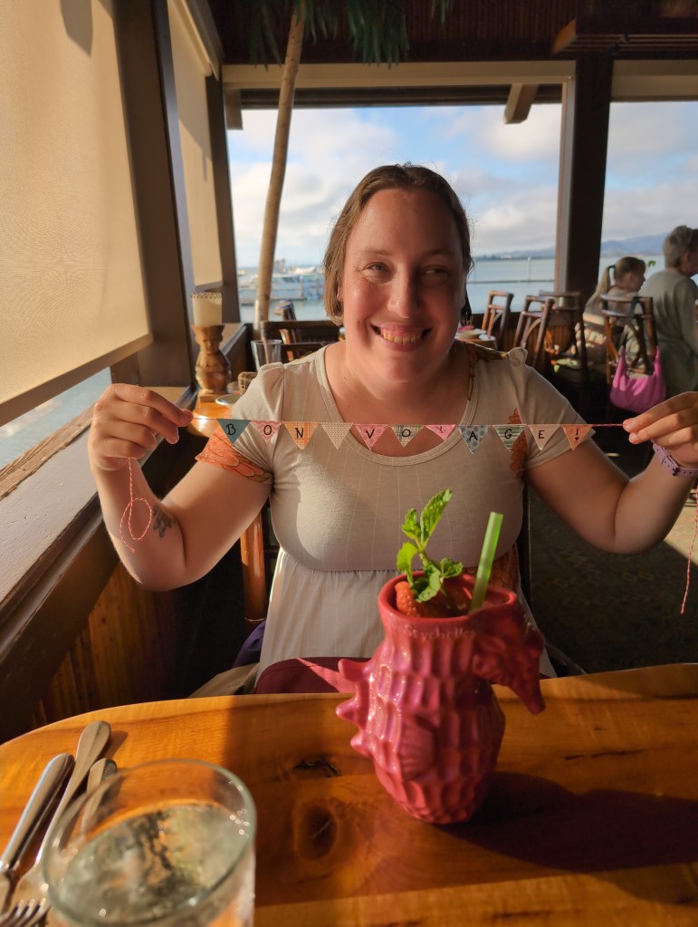 Tiffany sits at a restaurant table holding up a small banner with flags spelling on "Bon Voyage". In front of her a seahorse shaped, coral colored glass holds a tropical drink.