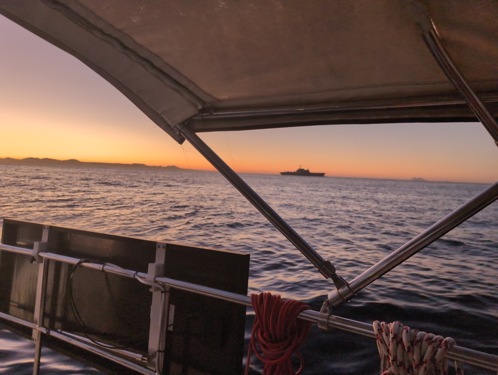 A large military vessel passes the viewers sailboat in the early light of morning.