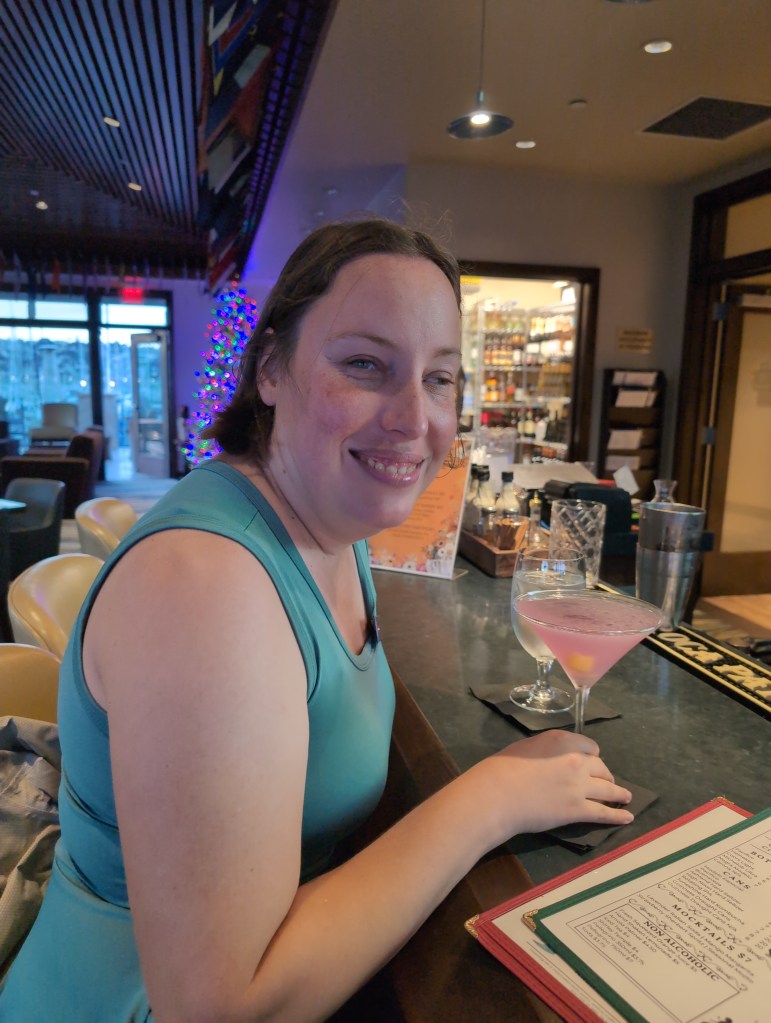 Tiffany sits at a bar, smiling at the camera, with a pink cocktail in front of her.