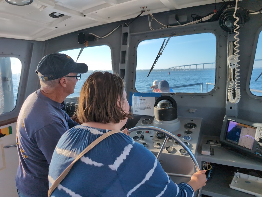 Tiffany, with a skipper beside her, steers a small military vessel toward a large bridge.