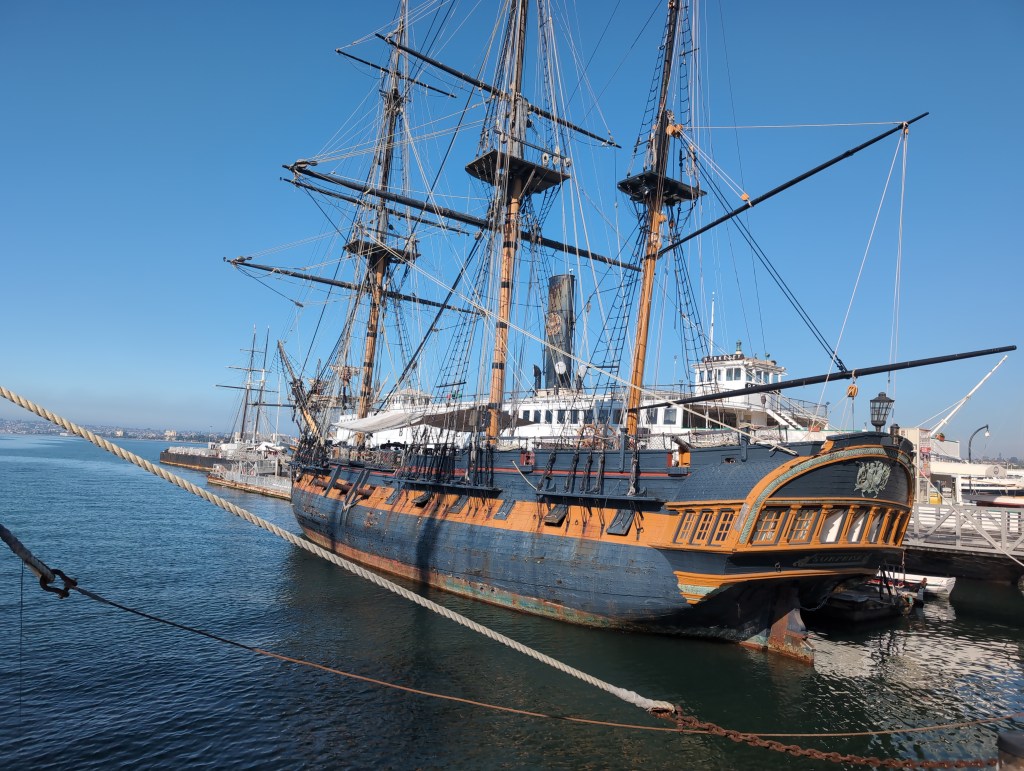 A three masted, square rigged, sailing frigate lies moored at a pier.