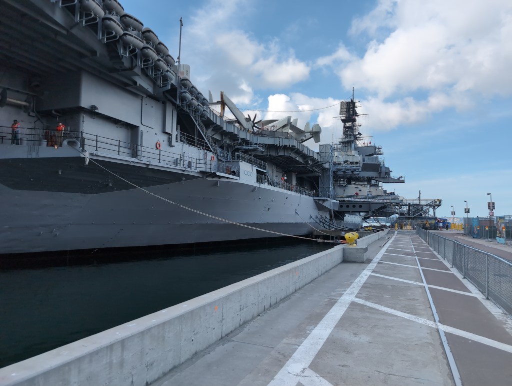 The starboard side of a massive aircraft carrier as view from a walkway beside the pier its moored at.