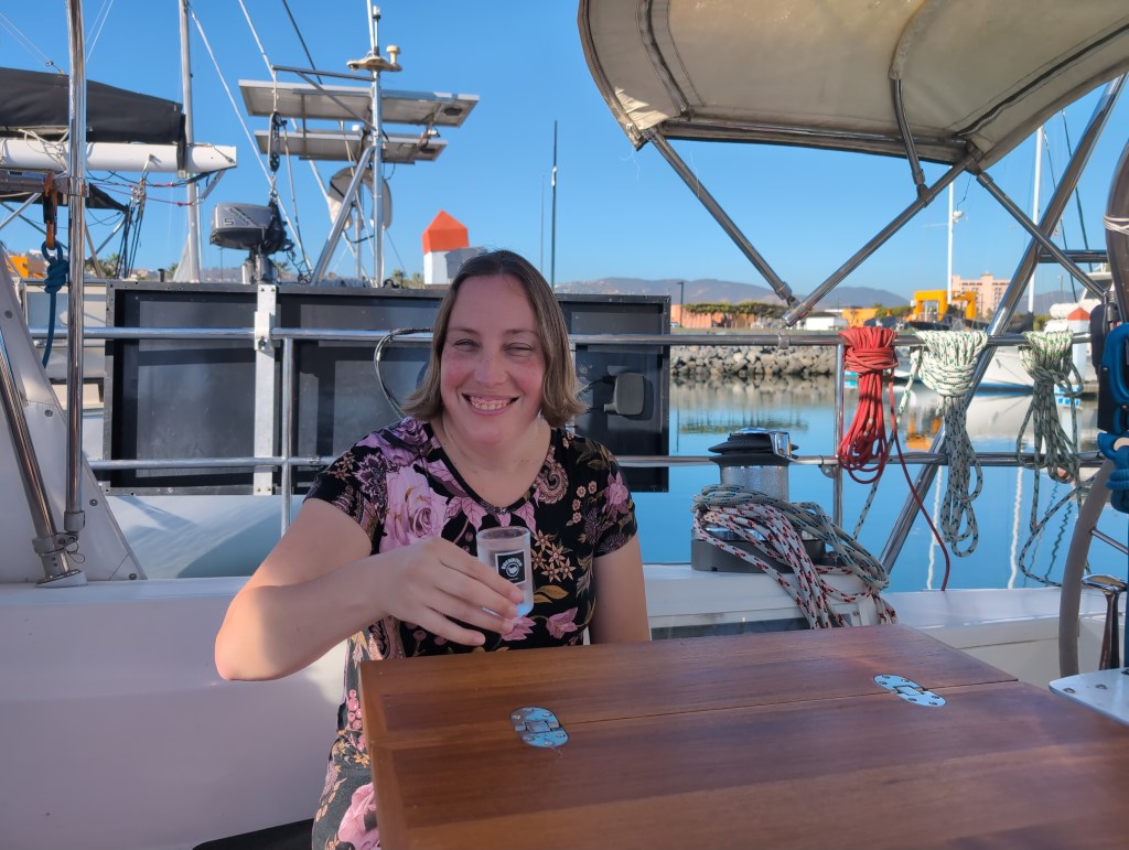 Tiffany, wearing a black shirt with pink flowers, sits before a wooden cockpit table, lifting a shot of clear liquid in a toast.