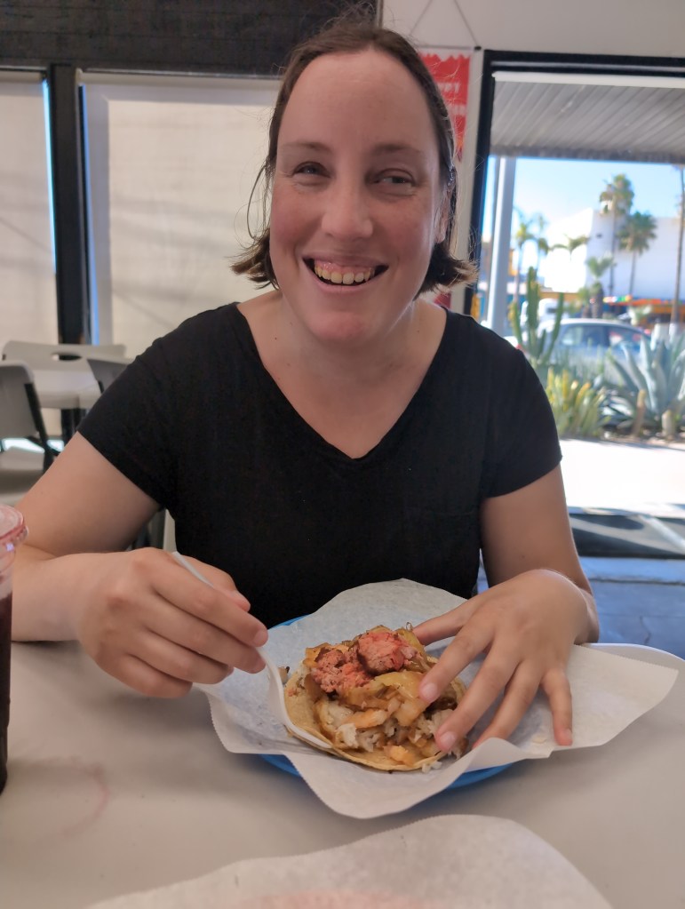 Tiffany, wearing a black V-necked t-shirt, smiles as she raises her fork over a tortilla mounded high with a mix of seafood.
